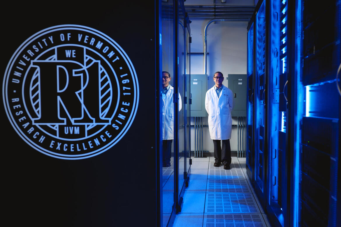 An individual in lab coat stands in a server room illuminated by blue light, with a University of Vermont research emblem prominently displayed on a black panel to the left.